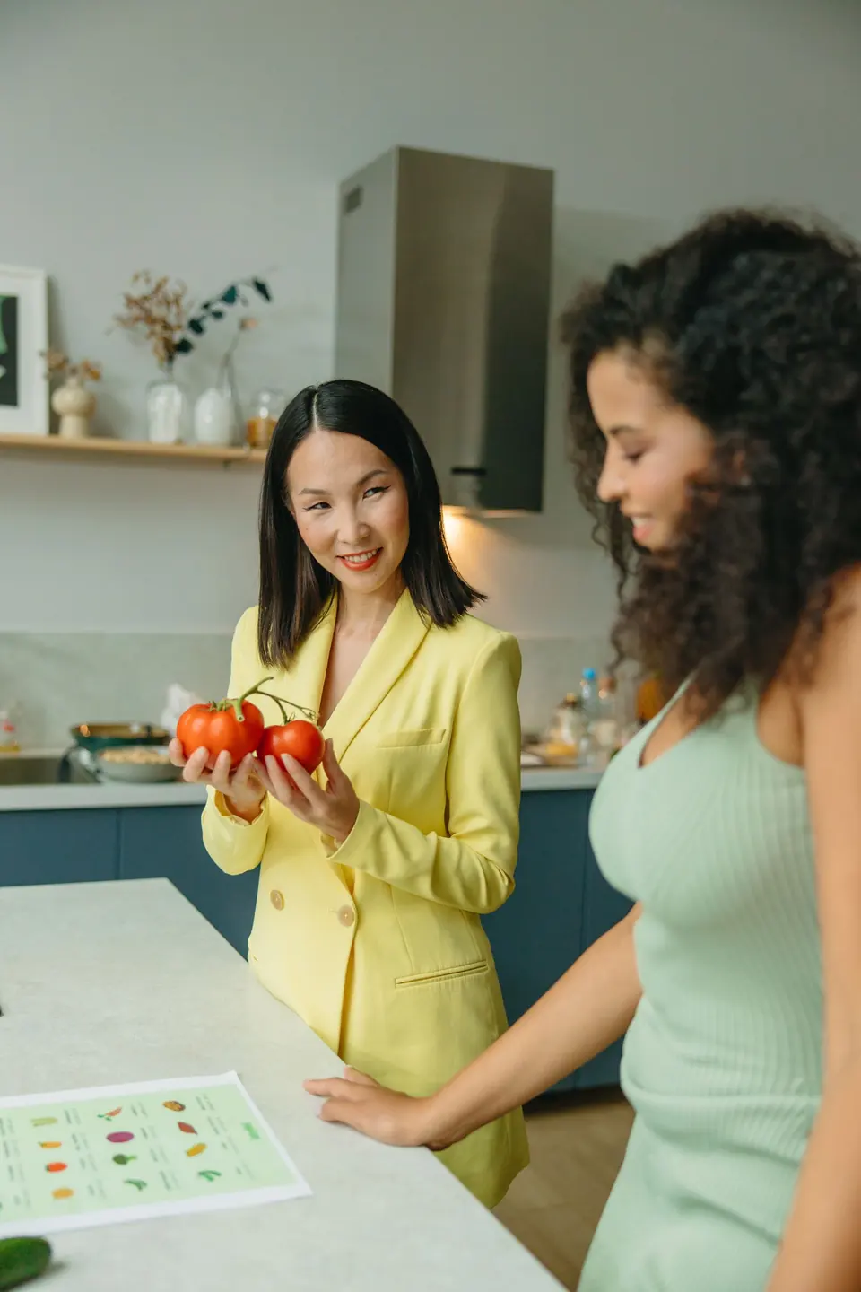 Diëtist Sanne in een geel pak geeft een cliënt in de keuken een tomaat